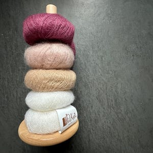 K&ouml;nnte beinhalten: Ein vertikaler Stapel aus f&uuml;nf flauschigen Garnkn&auml;ueln in den Farben Dunkelmagenta, Zartrosa, Beige und Wei&szlig;, gehalten von einem hellen Holzst&auml;nder. Das untere wei&szlig;e Garnkn&auml;uel tr&auml;gt ein wei&szlig;es Papieretikett mit der Aufschrift P&K in brauner Tinte.
