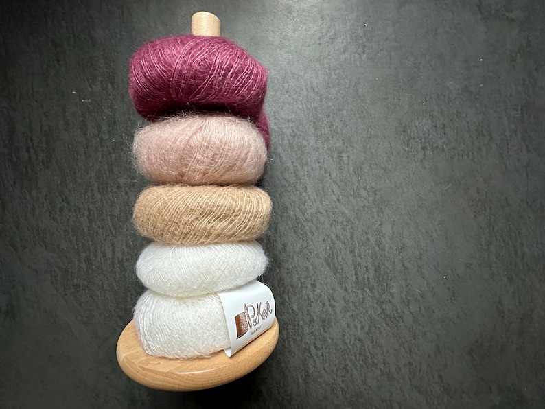 K&ouml;nnte beinhalten: Ein vertikaler Stapel aus f&uuml;nf flauschigen Garnkn&auml;ueln in den Farben Dunkelmagenta, Zartrosa, Beige und Wei&szlig;, gehalten von einem hellen Holzst&auml;nder. Das untere wei&szlig;e Garnkn&auml;uel tr&auml;gt ein wei&szlig;es Papieretikett mit der Aufschrift P&K in brauner Tinte.