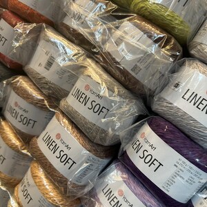 以下が含まれることがあります： さまざまな色のYarnArt Linen Soft糸の複数のスプールをクローズアップで撮影。スプールは透明なプラスチックで包装され、ブランド名と糸の種類が記載されています。オレンジ、茶色、緑、紫などの色があります。