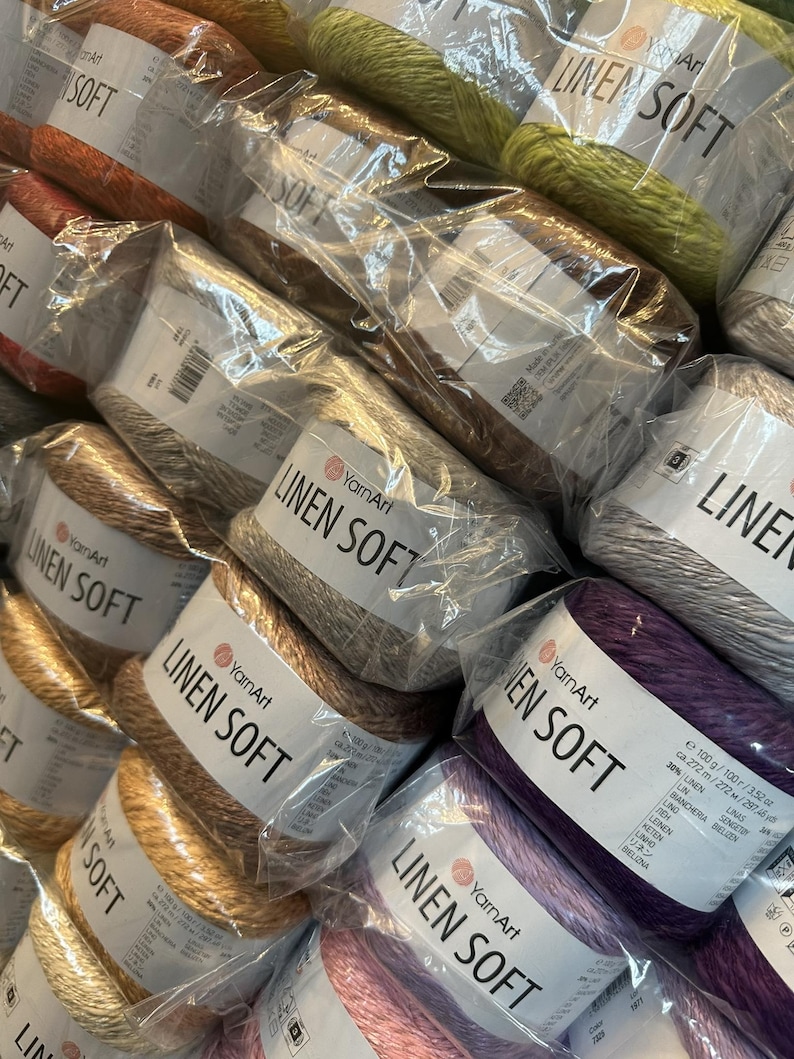 以下が含まれることがあります： さまざまな色のYarnArt Linen Soft糸の複数のスプールをクローズアップで撮影。スプールは透明なプラスチックで包装され、ブランド名と糸の種類が記載されています。オレンジ、茶色、緑、紫などの色があります。