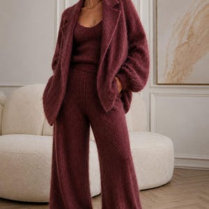 K&ouml;nnte beinhalten: Ein dreiteiliges Set in tiefem Burgunderrot, bestehend aus einem flauschigen, lang&auml;rmeligen offenen Blazer mit Revers, einem passenden Tanktop mit Rundhalsausschnitt und einer hoch taillierten Hose mit weitem Bein aus weichem Strickstoff.