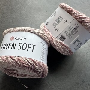 Puede incluir: Dos ovillos de hilo YarnArt Linen Soft en un color rosa claro y crema. La etiqueta dice "YarnArt Linen Soft" e incluye el n&uacute;mero de color 7405 y el n&uacute;mero de lote 1976. El hilo est&aacute; hecho en Turqu&iacute;a.