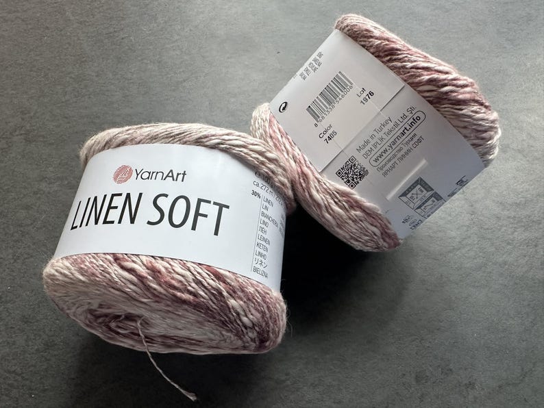 Puede incluir: Dos ovillos de hilo YarnArt Linen Soft en un color rosa claro y crema. La etiqueta dice "YarnArt Linen Soft" e incluye el n&uacute;mero de color 7405 y el n&uacute;mero de lote 1976. El hilo est&aacute; hecho en Turqu&iacute;a.