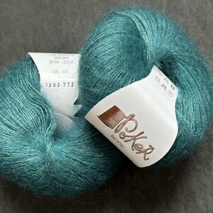 K&ouml;nnte beinhalten: Zwei Kn&auml;uel aus blaugr&uuml;nem Mohair- und Seidengarn mit flauschiger Textur. Jedes Kn&auml;uel hat ein wei&szlig;es Papieretikett mit dem Text Poker, Made in Italy, 25 gr - 212 m und 1563 773. Das Garn hat ein weiches, geb&uuml;rstetes Aussehen.