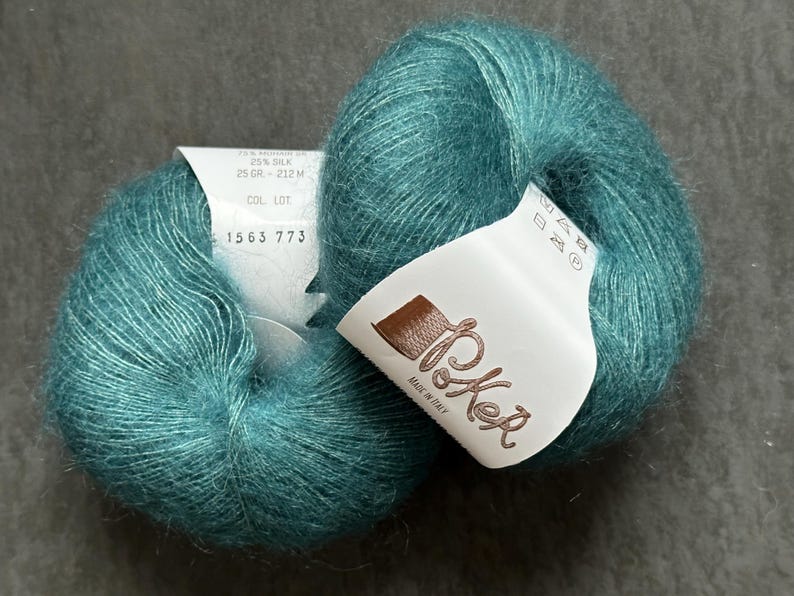 K&ouml;nnte beinhalten: Zwei Kn&auml;uel aus blaugr&uuml;nem Mohair- und Seidengarn mit flauschiger Textur. Jedes Kn&auml;uel hat ein wei&szlig;es Papieretikett mit dem Text Poker, Made in Italy, 25 gr - 212 m und 1563 773. Das Garn hat ein weiches, geb&uuml;rstetes Aussehen.