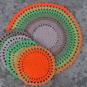 Puede incluir: Un conjunto de cuatro manteles individuales de ganchillo en tonos de verde, naranja y marr&oacute;n. Los manteles individuales est&aacute;n superpuestos, con el m&aacute;s grande en la parte inferior y el m&aacute;s peque&ntilde;o en la parte superior.