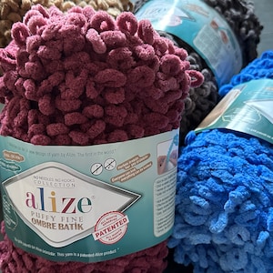 Może przedstawiać: Zbliżenie na kilka motków przędzy Alize Puffy Fine Ombre Batik w różnych kolorach, w tym bordowym, niebieskim, szarym i brązowym. Przędza ma pętelkową teksturę, a opakowanie zawiera napis "No Needles No Hooks Collection".