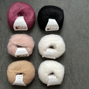 K&ouml;nnte beinhalten: Sechs runde Kn&auml;uel aus flauschigem Garn, angeordnet in zwei Spalten. Die Farben umfassen tiefes Magenta, Schwarz, zartes Rosa sowie drei Wei&szlig;- und Beiget&ouml;ne. Jedes Kn&auml;uel hat ein wei&szlig;es Papieretikett mit dem aufgedruckten Text Poker.