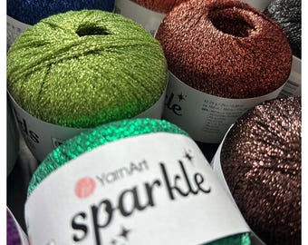 Op de afbeelding: Close-up van verschillende bollen YarnArt Sparkle garen in diverse kleuren, waaronder groen, koper en blauw. Het garen is metallic en glinsterend, perfect voor brei- en haakprojecten. Het etiket op de bol leest "Sparkle".