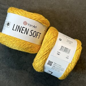 以下が含まれることがあります： 2つの黄色のYarnArt Linen Soft糸玉。糸は、ブランド名と製品情報が記載された白いラベルに巻かれています。糸は明るく、晴れた黄色です。