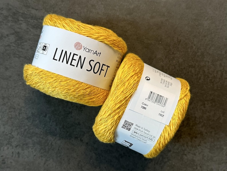 以下が含まれることがあります： 2つの黄色のYarnArt Linen Soft糸玉。糸は、ブランド名と製品情報が記載された白いラベルに巻かれています。糸は明るく、晴れた黄色です。