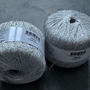 Op de afbeelding: Twee spoelen zilverkleurig garen met glitter en pailletten. Het garen is gelabeld "Parla C-5551-P-6 8 680131 072682".