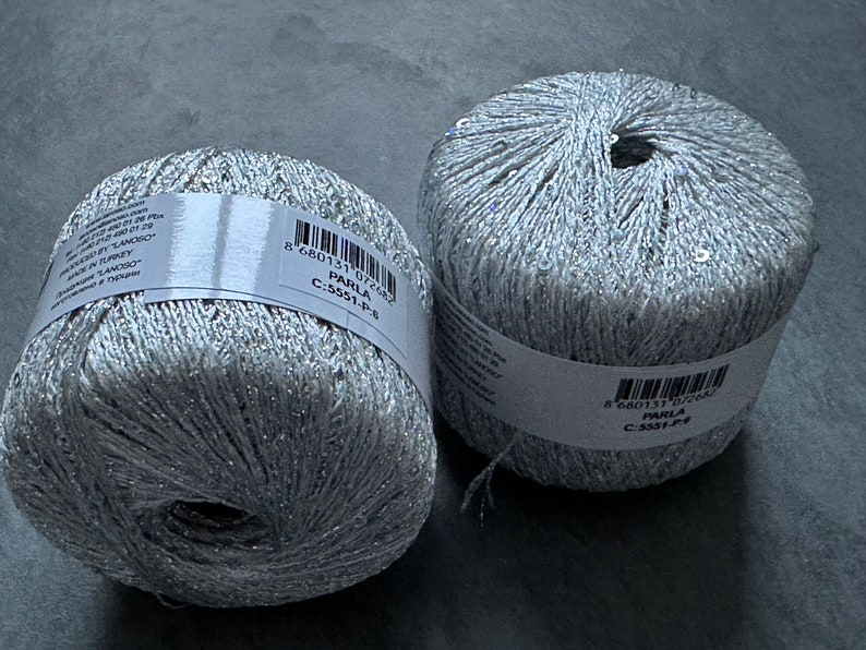 Op de afbeelding: Twee spoelen zilverkleurig garen met glitter en pailletten. Het garen is gelabeld "Parla C-5551-P-6 8 680131 072682".
