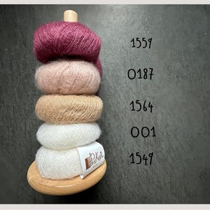 K&ouml;nnte beinhalten: Ein vertikaler Holzst&auml;nder mit f&uuml;nf gestapelten Kn&auml;ueln aus flauschigem Garn in den Farben Dunkelmagenta, Hellrosa, Beige und Wei&szlig;. Daneben stehen die Zahlen 1559, 0187, 1564, 001 und 1549. Ein wei&szlig;es Etikett am untersten Kn&auml;uel tr&auml;gt die Aufschrift PoKnit.