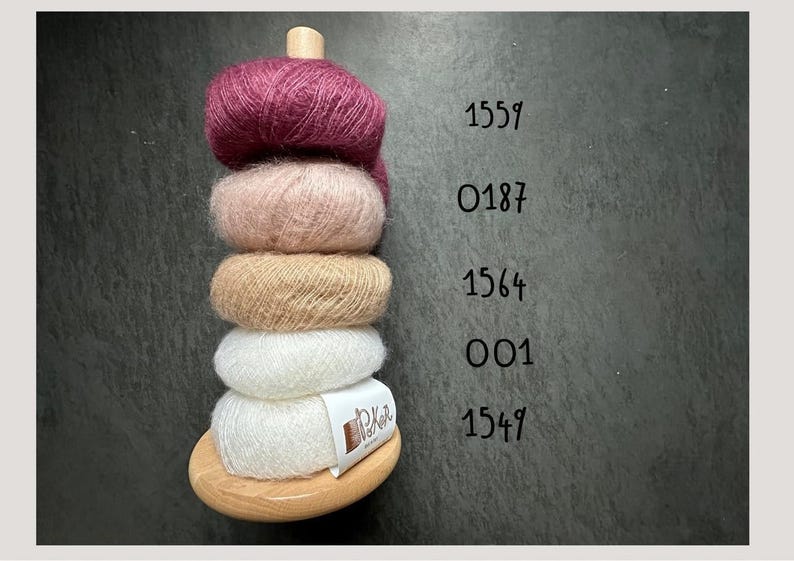 K&ouml;nnte beinhalten: Ein vertikaler Holzst&auml;nder mit f&uuml;nf gestapelten Kn&auml;ueln aus flauschigem Garn in den Farben Dunkelmagenta, Hellrosa, Beige und Wei&szlig;. Daneben stehen die Zahlen 1559, 0187, 1564, 001 und 1549. Ein wei&szlig;es Etikett am untersten Kn&auml;uel tr&auml;gt die Aufschrift PoKnit.