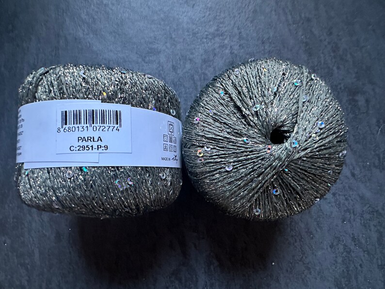 Op de afbeelding: Twee bollen zilver en grijs garen met iriserende pailletten. Het etiket vermeldt "PARLA C:2951-P-9 8 680131 072774"