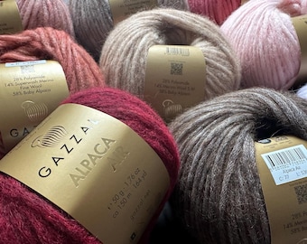 May include: A collection of Gazzal Alpaca Air yarn balls in various colours including deep red, brown, beige, and soft pink. Each ball features a gold paper label detailing a composition of 58% baby alpaca, 28% polyamide, and 14% merino wool, 50g, 1.76 oz.