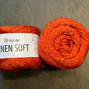 以下が含まれることがあります： 2つのオレンジ色の糸玉。1つは「YarnArt LINEN SOFT」の文字が入った白いラベルが付いています。糸は鮮やかなオレンジ色で、糸の質感がわかります。糸玉は灰色の表面にあります。