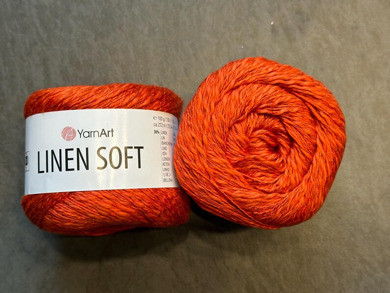 以下が含まれることがあります： 2つのオレンジ色の糸玉。1つは「YarnArt LINEN SOFT」の文字が入った白いラベルが付いています。糸は鮮やかなオレンジ色で、糸の質感がわかります。糸玉は灰色の表面にあります。