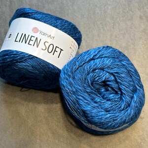 以下が含まれることがあります： 2つの青い糸玉。1つは「Linen Soft」の文字とYarnArtのロゴが入った白いラベルが付いています。糸は濃い青と薄い青の混色です。編み物やかぎ針編みのプロジェクトに適した糸です。