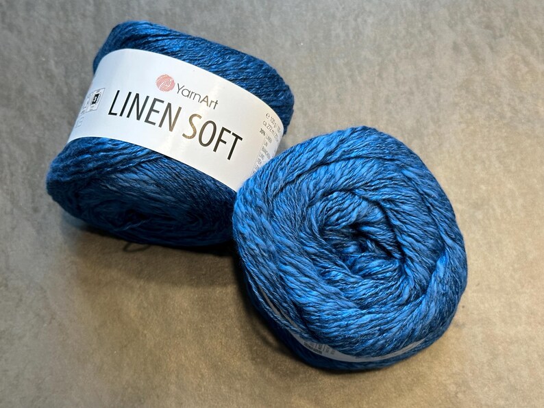 以下が含まれることがあります： 2つの青い糸玉。1つは「Linen Soft」の文字とYarnArtのロゴが入った白いラベルが付いています。糸は濃い青と薄い青の混色です。編み物やかぎ針編みのプロジェクトに適した糸です。