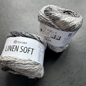 Puede incluir: Dos ovillos de hilo YarnArt Linen Soft. El hilo es una mezcla de lino y algod&oacute;n y es de color gris claro con vetas negras. El hilo est&aacute; etiquetado como "YarnArt Linen Soft" y tiene instrucciones de cuidado en la etiqueta.