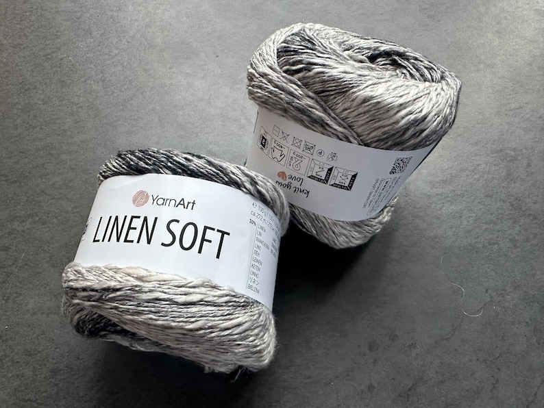 Puede incluir: Dos ovillos de hilo YarnArt Linen Soft. El hilo es una mezcla de lino y algod&oacute;n y es de color gris claro con vetas negras. El hilo est&aacute; etiquetado como "YarnArt Linen Soft" y tiene instrucciones de cuidado en la etiqueta.