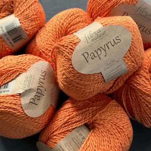 Könnte beinhalten: Eine Ansammlung von orangefarbenen Garnknäueln, jedes mit einem Etikett "fibra natura Papyrus". Das Garn ist wahrscheinlich eine Baumwollmischung, geeignet für verschiedene Strick- und Häkelprojekte. Die Knäuel werden in Gramm gemessen.