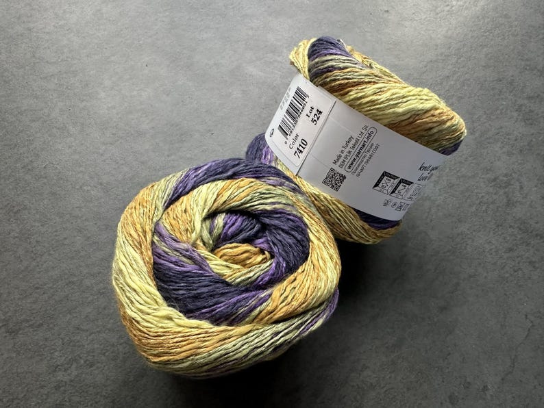 Puede incluir: Dos ovillos de hilo en una combinaci&oacute;n de colores amarillo, morado y verde. El hilo est&aacute; etiquetado con "Color 7410 Lot 324".