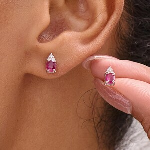 Könnte beinhalten: Kleine, tropfenförmige Ohrringe mit einem leuchtend rosa Edelstein an der Basis und einem kleinen, dreieckigen weißen Stein an der Spitze. Die Ohrringe sind in Silber oder weißem Metall gefasst. Ein Ohrring wird ans Ohr gehalten.