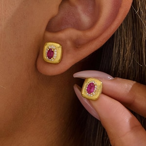 Könnte beinhalten: Goldfarbene quadratische Ohrstecker mit einem ovalen Rubin in der Mitte, umgeben von kleinen weißen Edelsteinen. Ein Ohrring wird ans Ohr gehalten, um das Design zu zeigen. Die Ohrringe sind vor einem hautfarbenen Hintergrund.