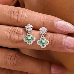 Könnte beinhalten: Ein Paar silberne Ohrringe mit floralem Design. Jeder Ohrring hat eine kleine Blume oben und eine vierblättrige Kleeblattform mit grünen Edelsteinen und weißen Akzenten. Die Ohrringe werden vor einem Hautton-Hintergrund gezeigt.