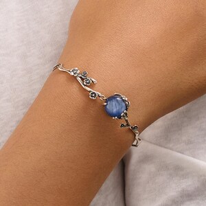 Puede incluir: Una pulsera de tono plateado con una piedra preciosa azul redonda en el centro. La banda presenta un motivo texturizado similar a una rama que se extiende desde la piedra, incorporando pequeños detalles florales u hojas oscuras en la cadena.