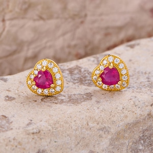 Puede incluir: Un par de pendientes de botón en forma de corazón. Cada pendiente presenta un centro de piedra preciosa roja vibrante, rodeado por un halo de piedras preciosas claras más pequeñas. El engaste es dorado, lo que añade un toque de elegancia. Los pendientes se muestran sobre una superficie texturizada de tono neutro.