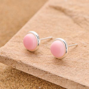 May include: Two stud earrings featuring a pale pink, circular stone set within a silver-coloured bezel. The earrings are presented on a light brown, textured surface. The design of the earrings is simple and minimalist.