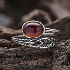 May include: A silver-toned bypass ring featuring an oval-shaped, deep red cabochon stone. The band is crafted with an oxidized finish and detailed with a textured, etched feather or leaf pattern that wraps around the finger for a distinct look.