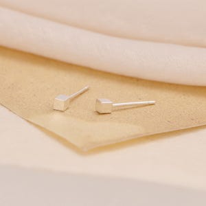 May include: A pair of minimalist silver stud earrings. Each earring features a small, square cube at the end of a thin post. The earrings are displayed on a tan surface with a soft, off-white fabric in the background.
