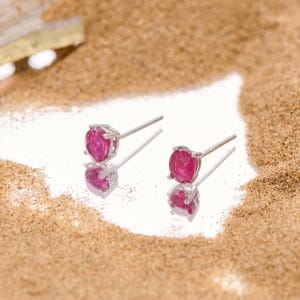 May include: A pair of silver stud earrings with round, vibrant pink gemstones. The earrings are set against a reflective surface, with a backdrop of sand. The gemstones are the focal point, showcasing their color and shape.