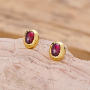 May include: A pair of gold stud earrings, each with a deep red, oval-shaped stone. The earrings are set on a light-coloured, textured surface, with a blurred wooden background.