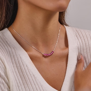 Könnte beinhalten: Eine silberne Halskette mit einer geschwungenen Reihe leuchtend rosa Edelsteine. Die Halskette ist zart und liegt anmutig am Hals. Die Edelsteine haben eine helle, auffällige Farbe und verleihen Eleganz.