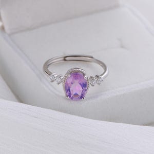 May include: A silver ring featuring a large oval-cut, light purple gemstone. The ring is accented with smaller, clear gemstones on either side of the main stone. The ring is set against a white background.