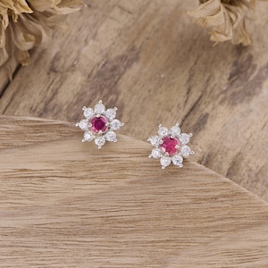 Könnte beinhalten: Ein Paar silberfarbene Ohrstecker. Jeder Ohrstecker hat einen zentralen, runden, tiefrosa Edelstein, der von kleineren, klaren Edelsteinen umgeben ist und ein blütenähnliches Design bildet. Die Ohrringe sind auf einem Holzgrund platziert.