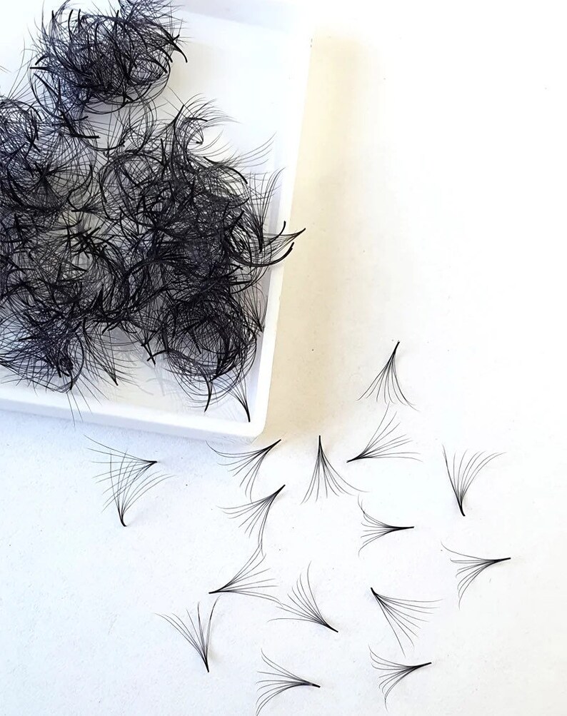 May include: A white tray with a pile of black eyelash extensions. Several individual extensions are scattered on a white surface.