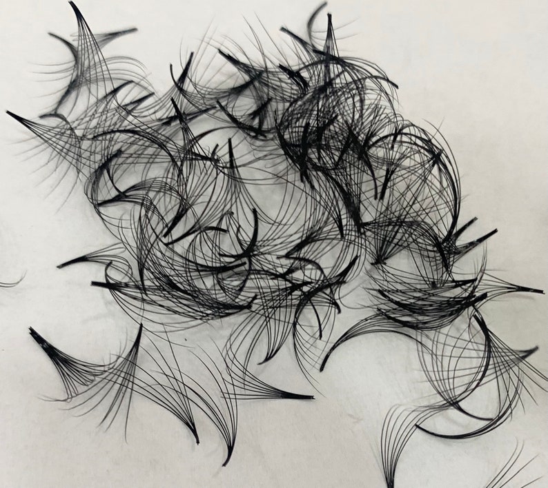 May include: A close-up of a pile of black individual eyelash extensions. The extensions are arranged in a random pattern on a white surface.