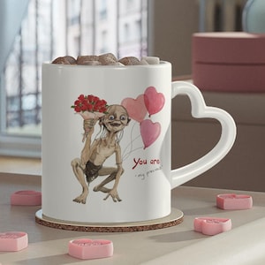 May include: White ceramic mug with a heart-shaped handle, featuring a cartoon character holding flowers and heart-shaped balloons. The mug has the text "You are my precious". The mug is on a wooden coaster, surrounded by pink heart-shaped candies.