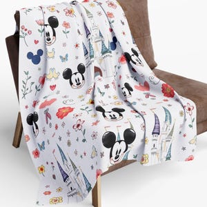 Può includere: Una coperta bianca con un motivo ripetuto di Topolino, castelli, fiori e farfalle multicolori, drappeggiata su una sedia marrone. La coperta è realizzata in materiale morbido.