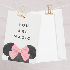 Peut inclure: Une carte blanche avec le texte "YOU ARE MAGIC" et un design inspiré de Minnie Mouse, avec des oreilles noires et un nœud rose. La carte est maintenue par une pince dorée à côté d'une enveloppe blanche.