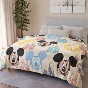 Può includere: Una coperta beige con un motivo ripetuto di facce di Topolino e castelli colorati. La coperta è su un letto con cuscini bianchi e una testiera grigia. Il design include il volto di Topolino in nero, blu e giallo.