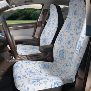 May include: Car seat covers featuring a blue and white toile pattern. The design includes illustrations of figures, bows, and floral elements. The covers are installed in a car, with the steering wheel and dashboard visible.