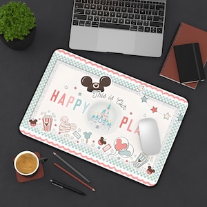 May include: A rectangular mousepad with a whimsical design featuring a castle, Mickey Mouse ears, and the words "HAPPY PLACE." The border is a light pink and mint green checkered pattern. A white mouse and a laptop are also visible.